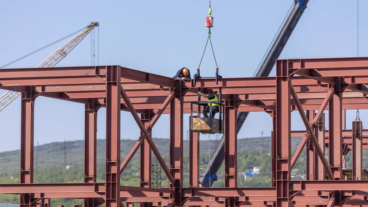 Workers assembling a red steel building frame with cranes overhead