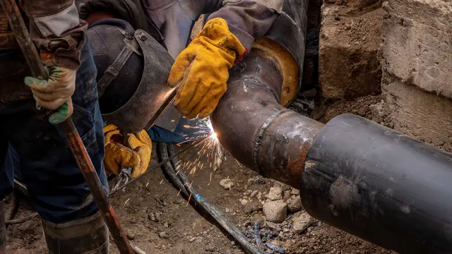 A welder in yellow gloves welding a steel pipe joint outdoors