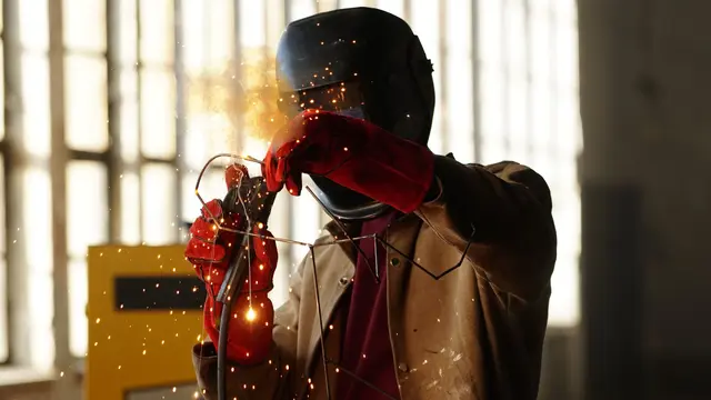 A welder in a leather jacket and red gloves welding beside a factory window