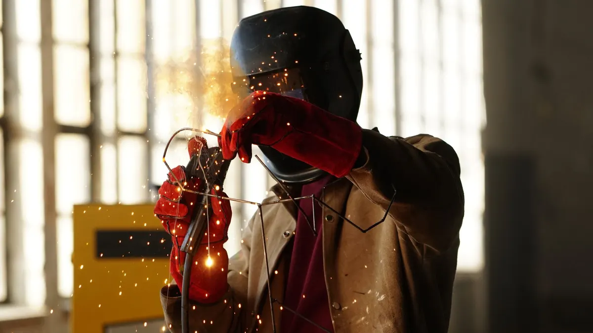 A welder in a leather jacket and red gloves welding beside a factory window