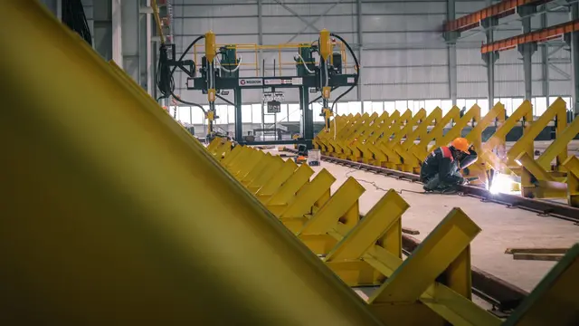 A welder working among long yellow steel frames in a fabrication warehouse
