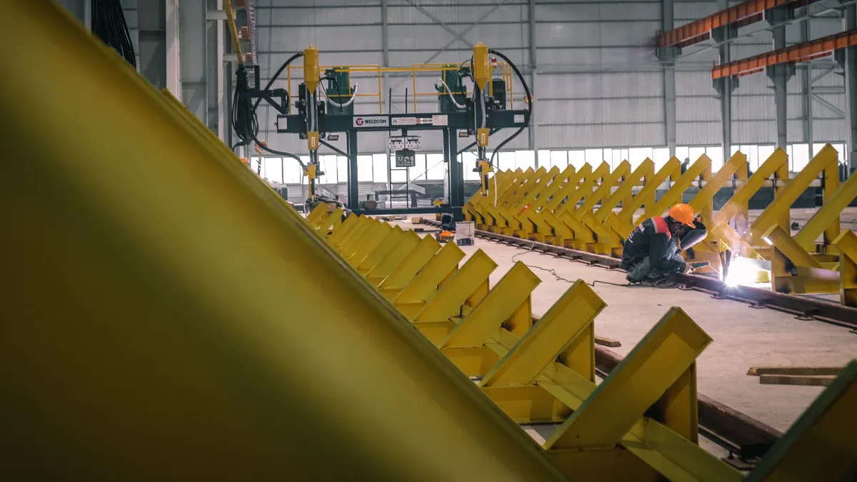 A welder working among long yellow steel frames in a fabrication warehouse