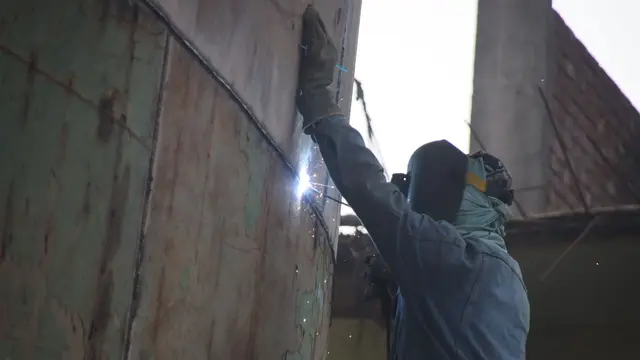 A welder standing against a ship's hull, welding a seam at a shipyard