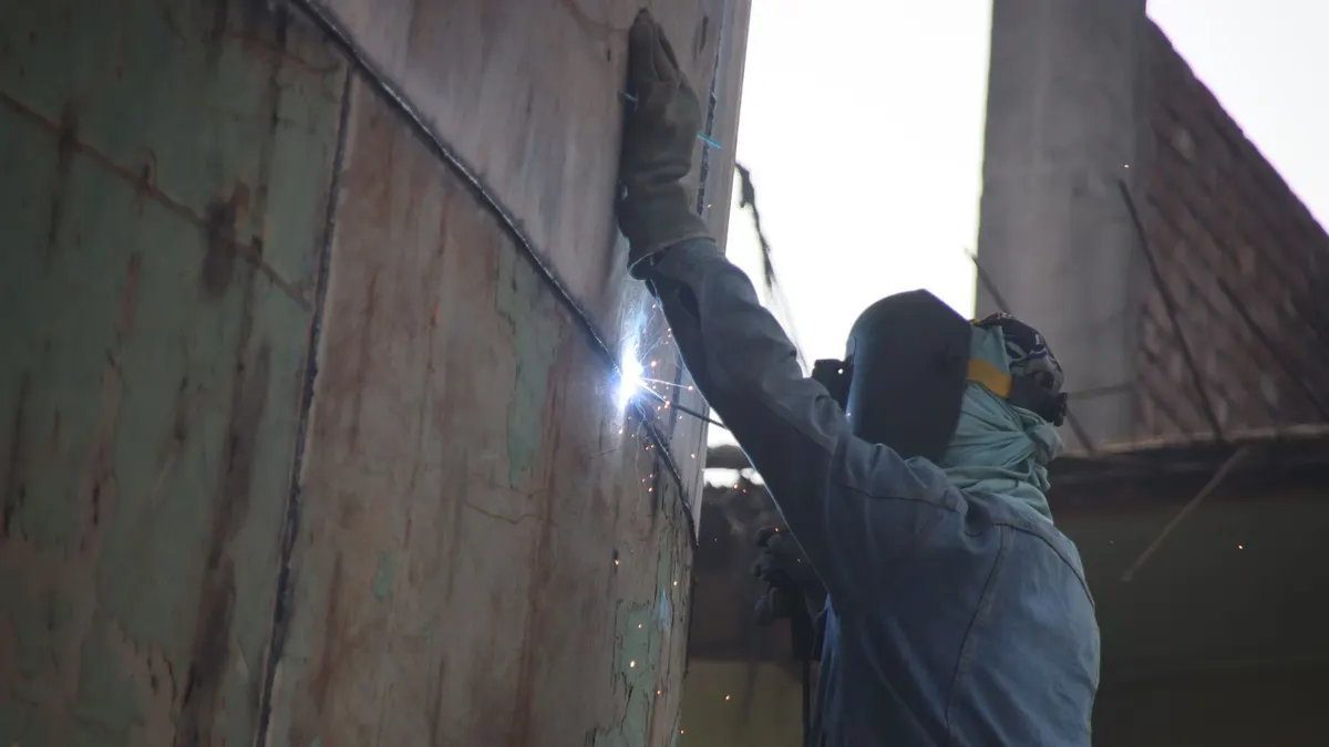 A welder standing against a ship's hull, welding a seam at a shipyard