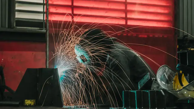 A welder in a helmet and blue shirt welding at a workstation