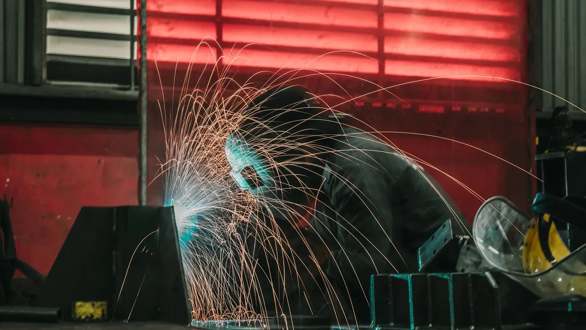 A welder in a helmet and blue shirt welding at a workstation