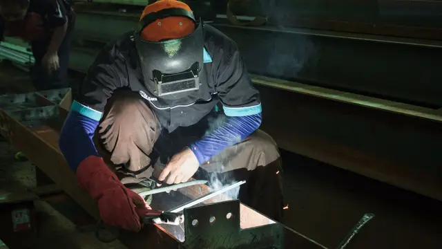 A welder in an orange hard hat crouched over a weld in a workshop