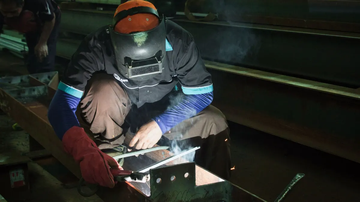 A welder in an orange hard hat crouched over a weld in a workshop