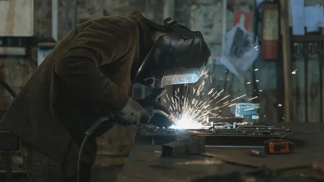 A welder at a workbench welding a metal piece with sparks flying