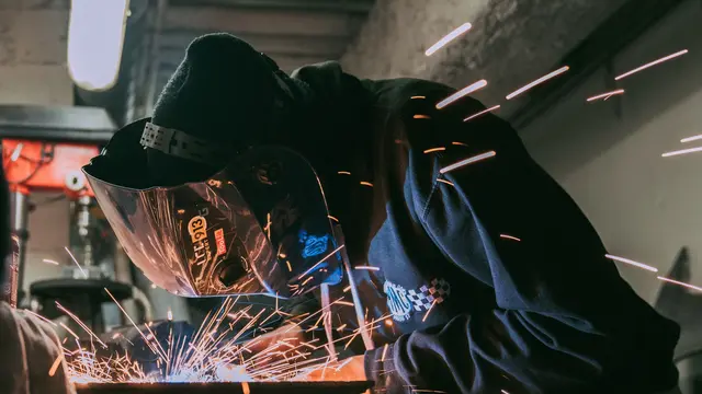 A welder in a cap and helmet with sparks flying across the workbench