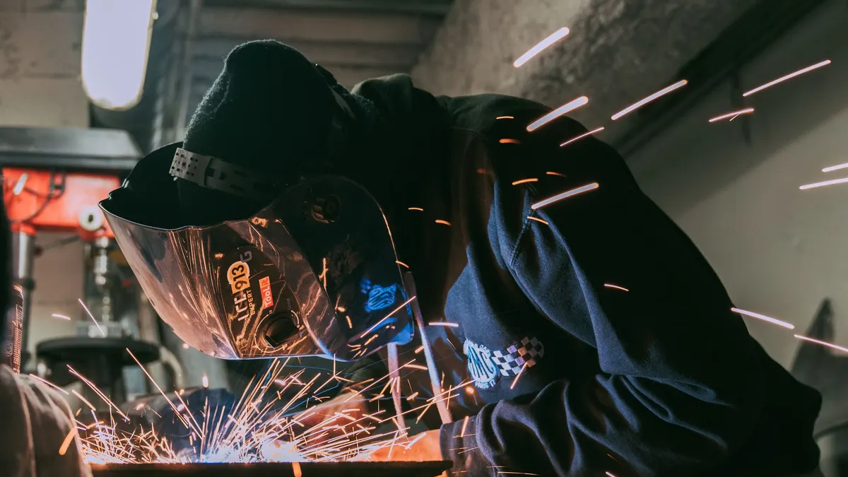 A welder in a cap and helmet with sparks flying across the workbench