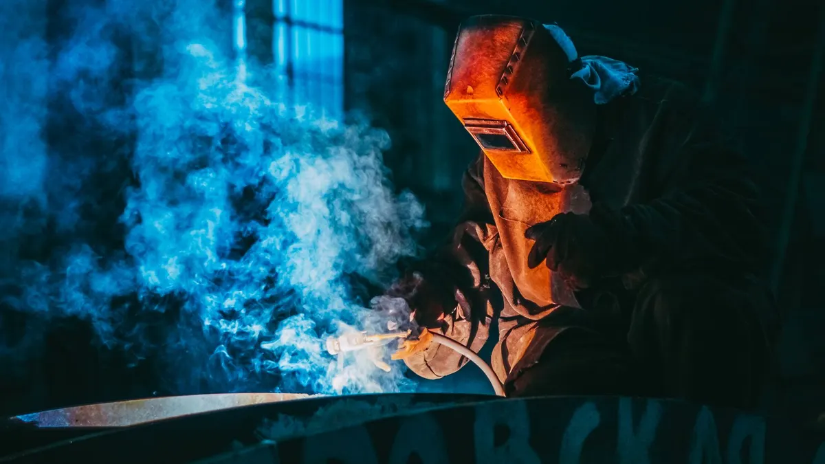 A welder in a helmet working through blue smoke in a dark workshop