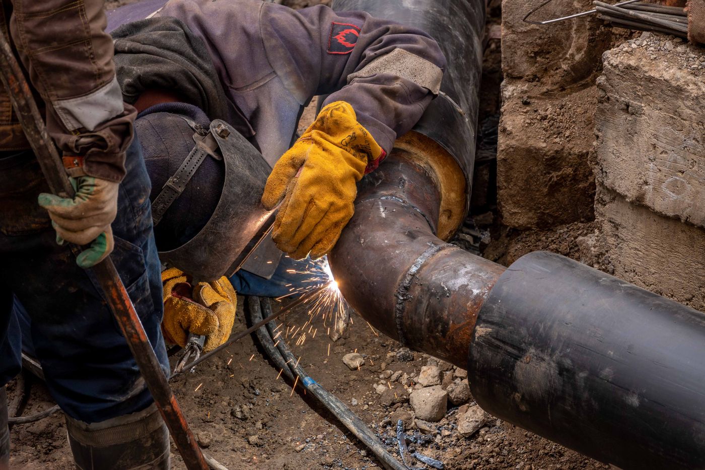 A welder in yellow gloves stick-welding a joint on a large steel pipe as sparks fall