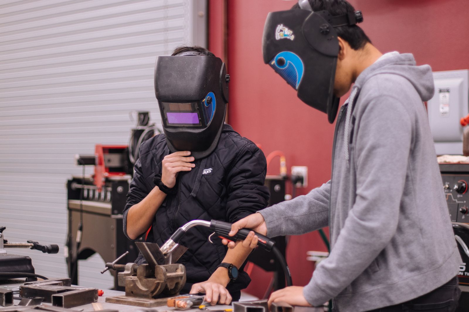 Two welding students in helmets at a workbench, one guiding a MIG torch