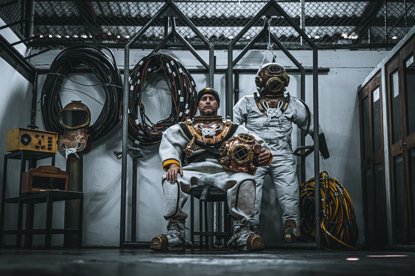 Two men in heavy commercial diving suits and helmets standing with their equipment