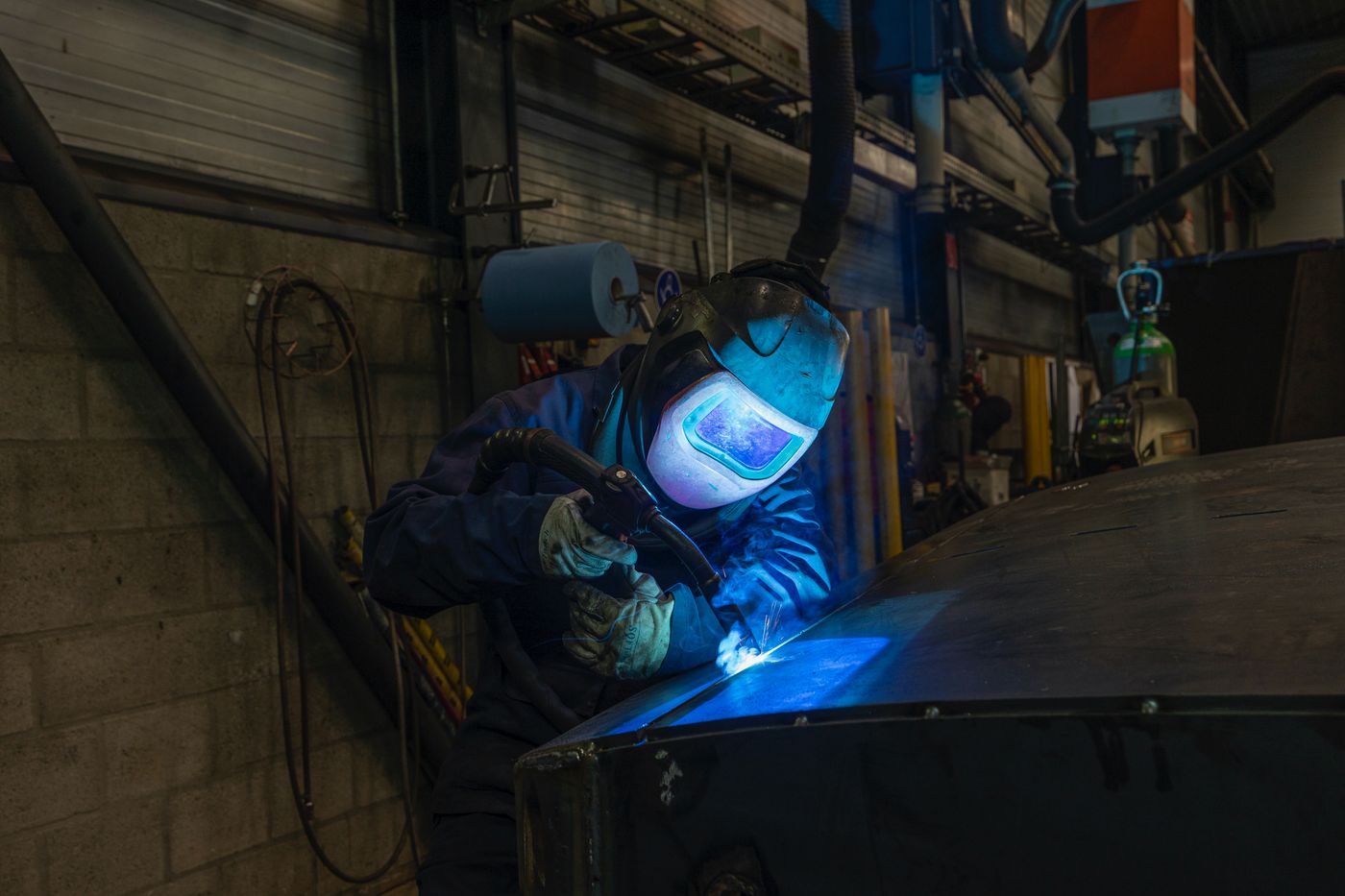 A welder in an auto-darkening helmet running a MIG bead on steel plate in a fabrication shop