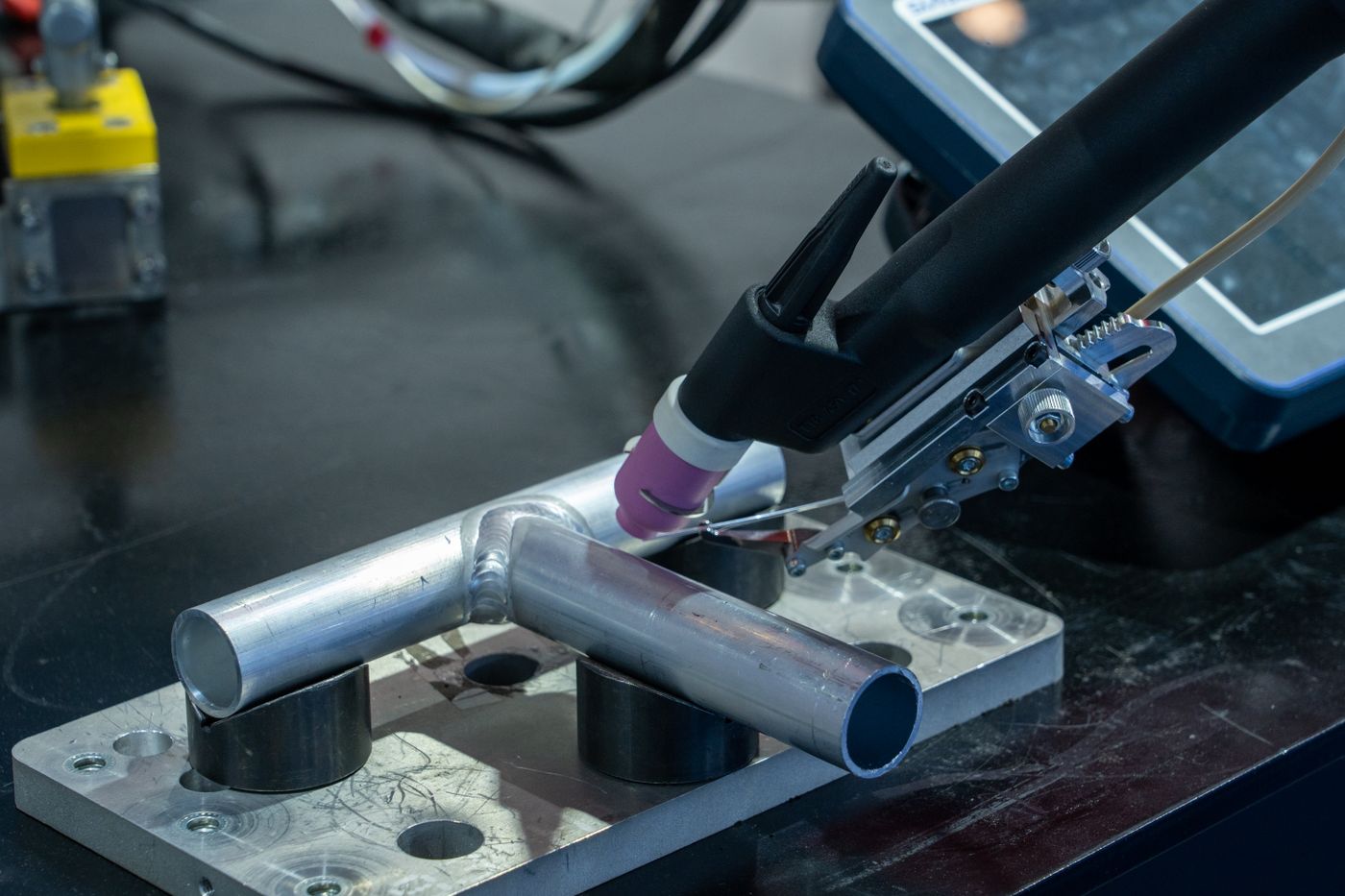 A TIG torch held in a fixture over an aluminum tube joint on a welding bench